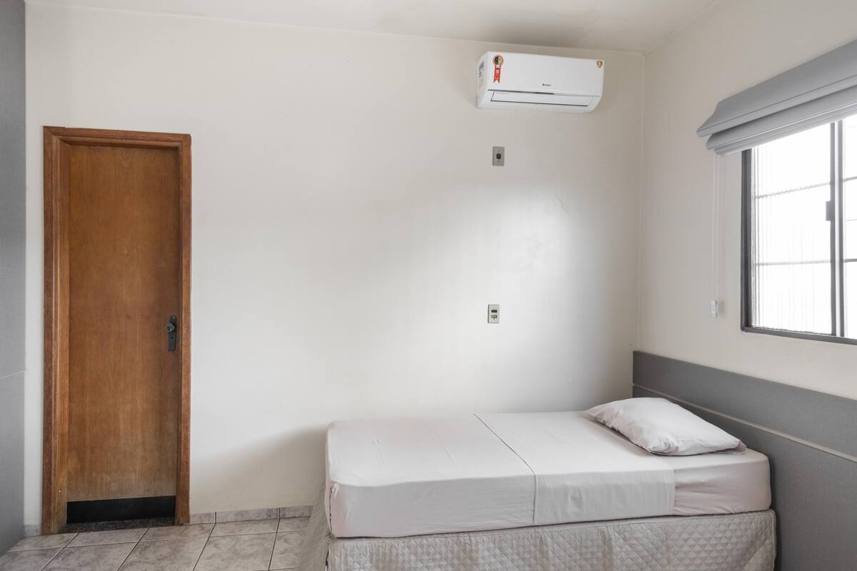 A small, single-bed room with a wooden door, a window with blinds, and a wall-mounted air conditioner.