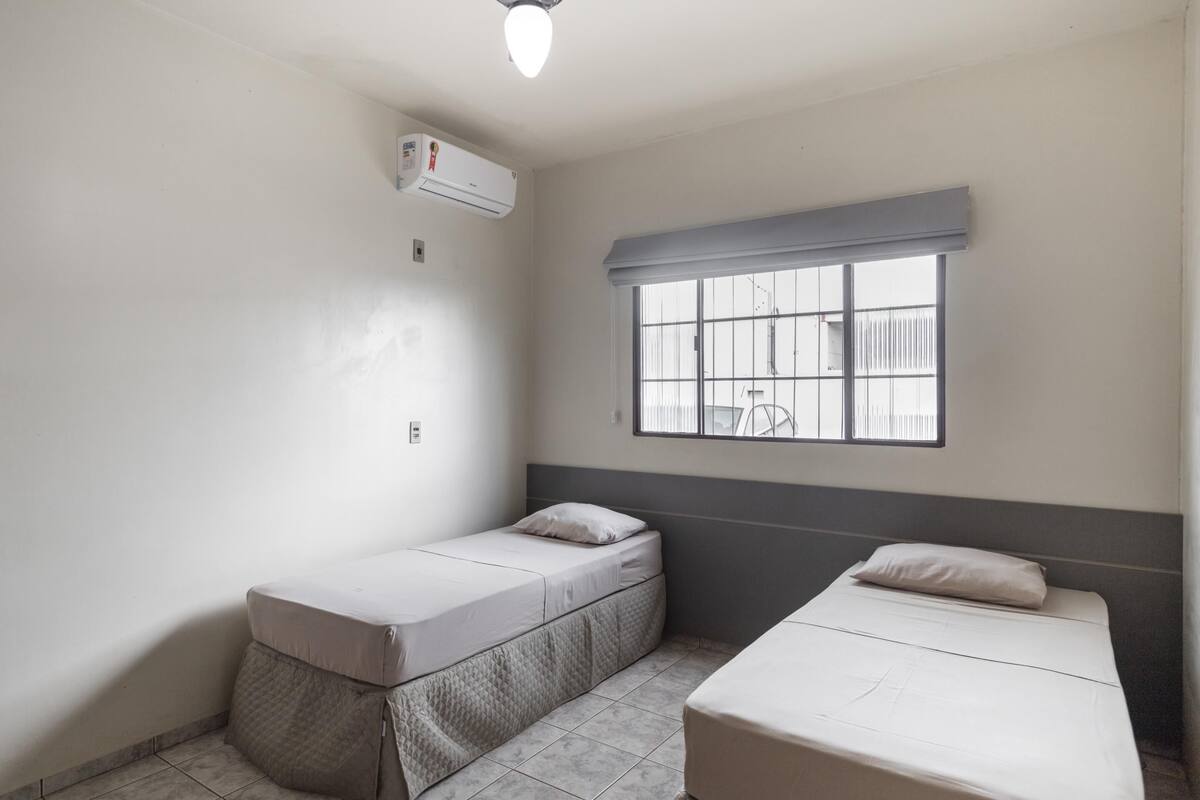 A small, clean room with two single beds, a window with bars, and an air conditioning unit.