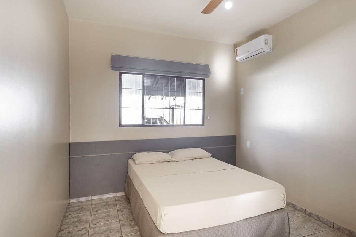 A bedroom with a bed, a window with blinds, an air conditioning unit, and a ceiling fan.