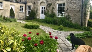 A stone building with a garden in front, a paved pathway, and a black dog standing on the path. - 30% Discount Beautiful converted studio apartment in idyllic Cotswold village! (Cirencester)