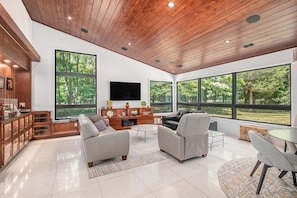 A spacious living area with a wooden ceiling, large windows, a flat-screen TV, and modern furniture.