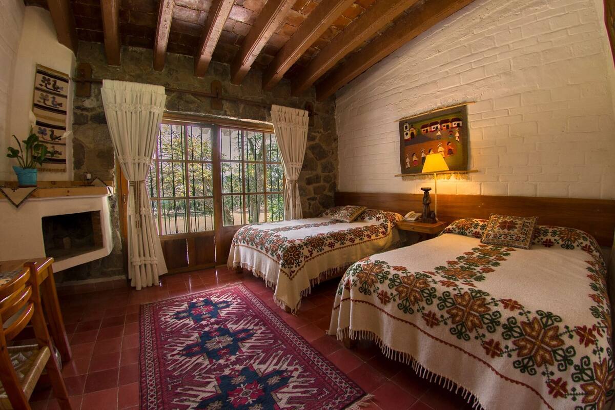Una habitación con dos camas, una chimenea, una ventana con cortinas, una mesita de noche y una lámpara fijada a la pared.