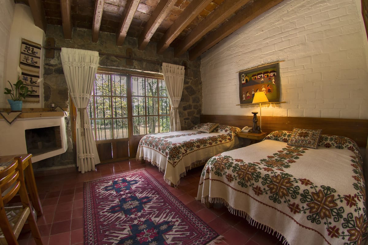 Una habitación con dos camas, una pared de piedra, una ventana con cortinas, un techo de madera y una mesita de noche con lámpara.