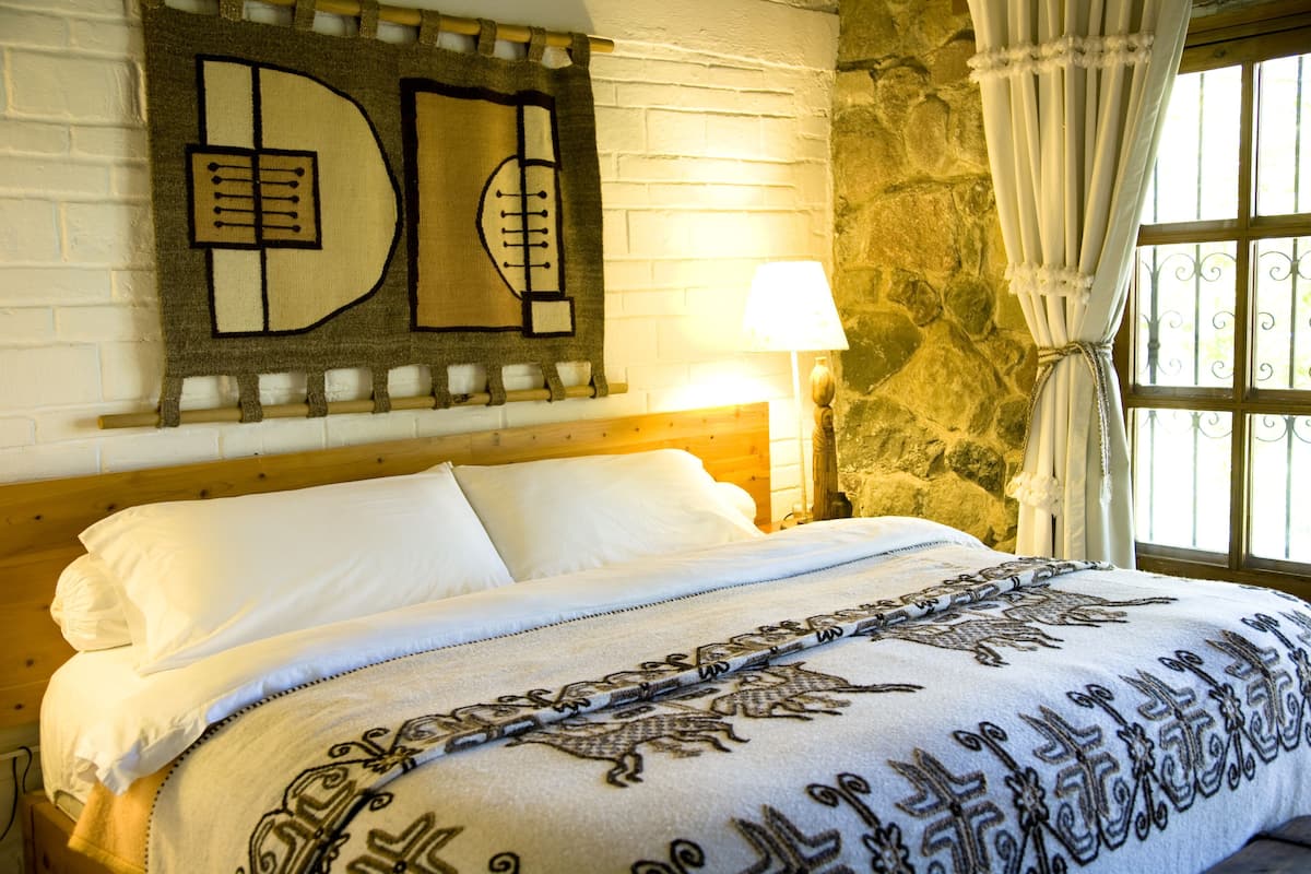 Una cama con sábanas blancas, una colcha estampada, una obra de arte en la pared, una lámpara y una pared de piedra.