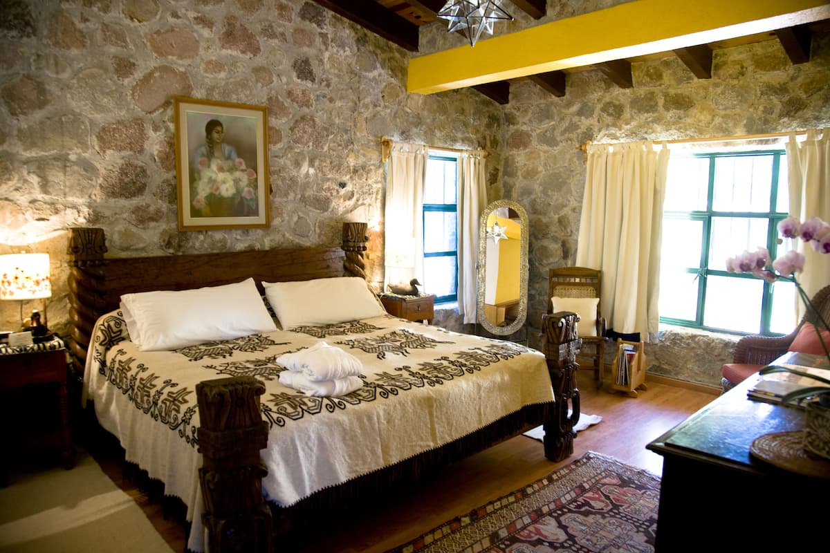Un dormitorio con paredes de piedra, una cama grande, cabecero de madera, mesita de noche con lámpara y un espejo en la pared.