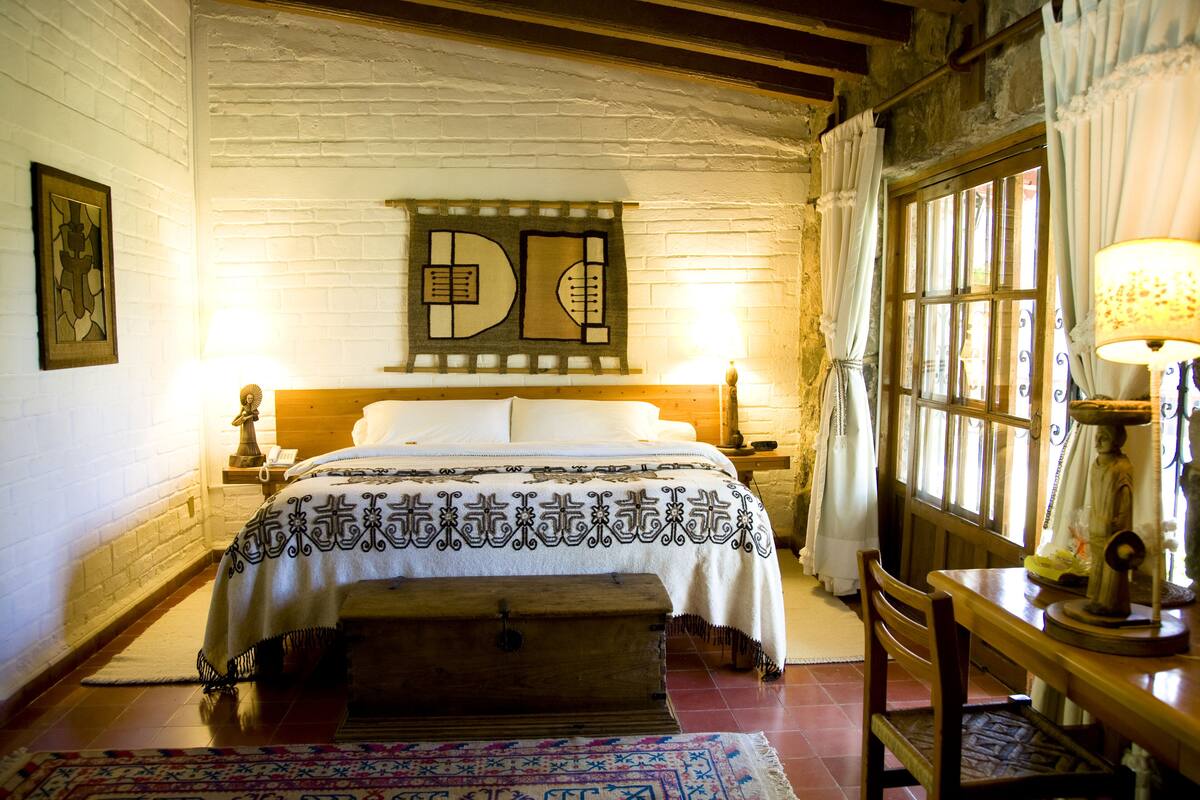 Una habitación tradicional con una cama, un cofre de madera, una mesa con una lámpara y un ventanal con cortinas.