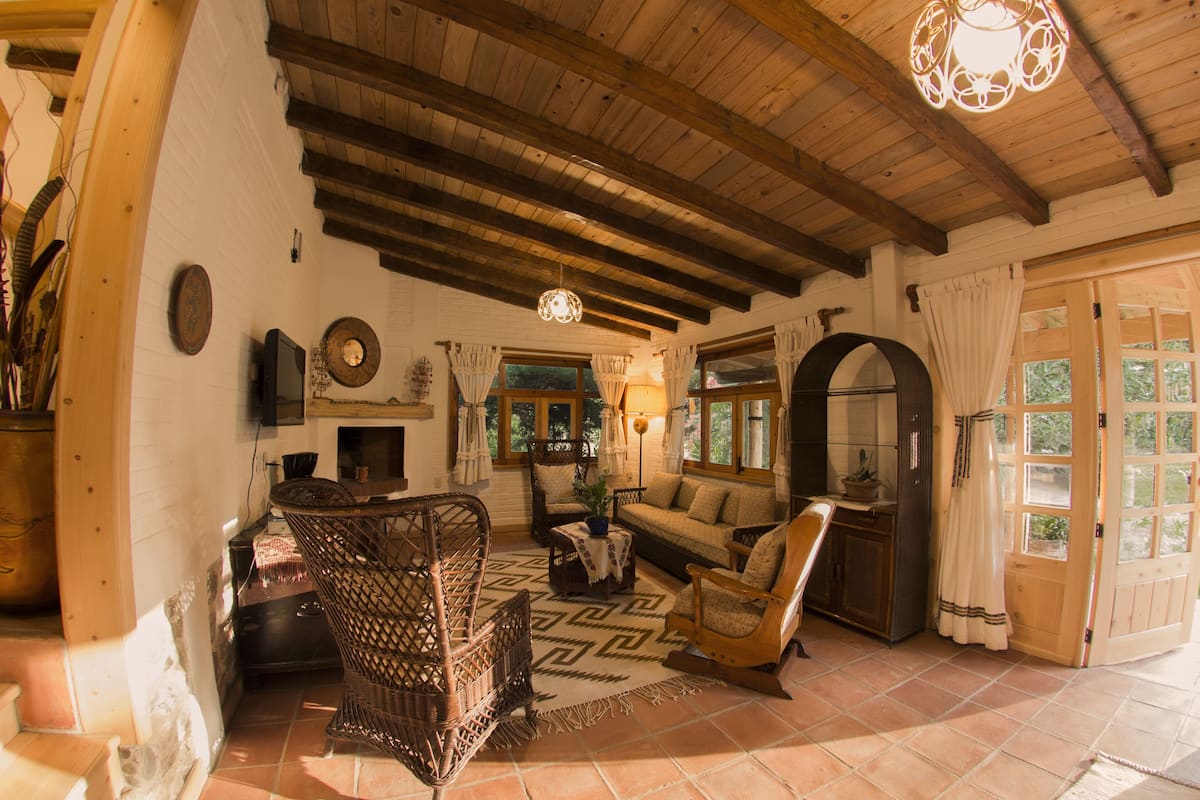 Una sala rústica con vigas de madera a la vista, una chimenea y una zona de estar acogedora con mobiliario de mimbre.
