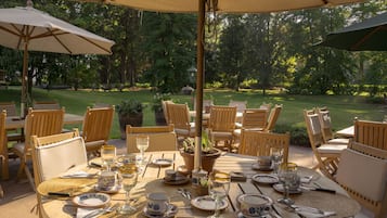 An outdoor dining area with tables set for a meal, surrounded by chairs and umbrellas, with a garden view.