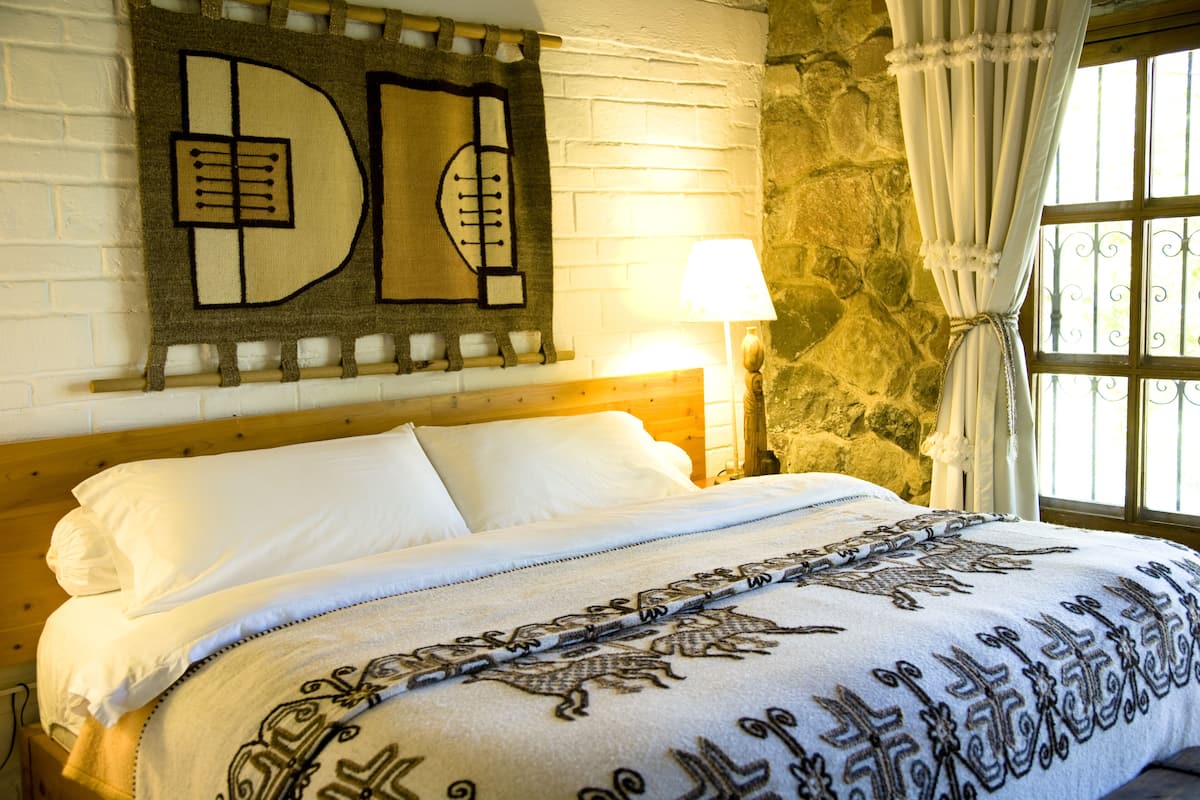 Una cama con una colcha estampada, una obra de arte colgada en la pared, una lámpara y una pared de piedra.