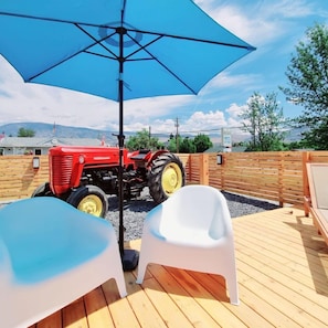 Eine Holzterrasse mit einem roten Traktor, zwei weißen Plastikstühlen und einem blauen Sonnenschirm.