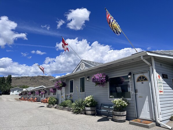 A row of single-story cabins with outdoor seating, a flagpole with flags, and a sign indicating the number of rooms. - The Orchard at Oliver (Oliver)