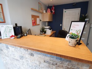 A reception desk with a stone wall, a bulletin board, flags, a security camera, and a sign about booking online. - The Orchard at Oliver (Oliver)