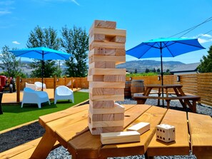 Un jeu de construction en bois sur une table de pique-nique, avec un dé et deux parasols bleus en arrière-plan.