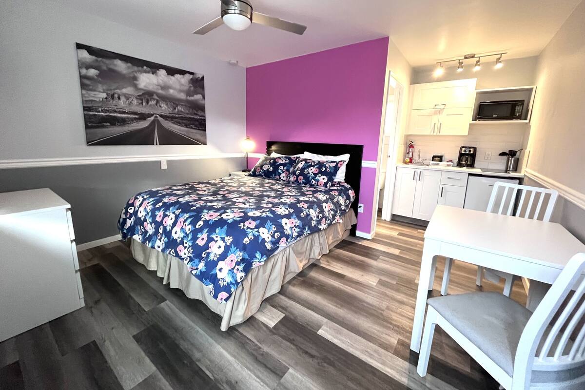 Un dormitorio con cama, escritorio, silla, ventilador de techo, una zona de cocina y un cuadro en la pared.