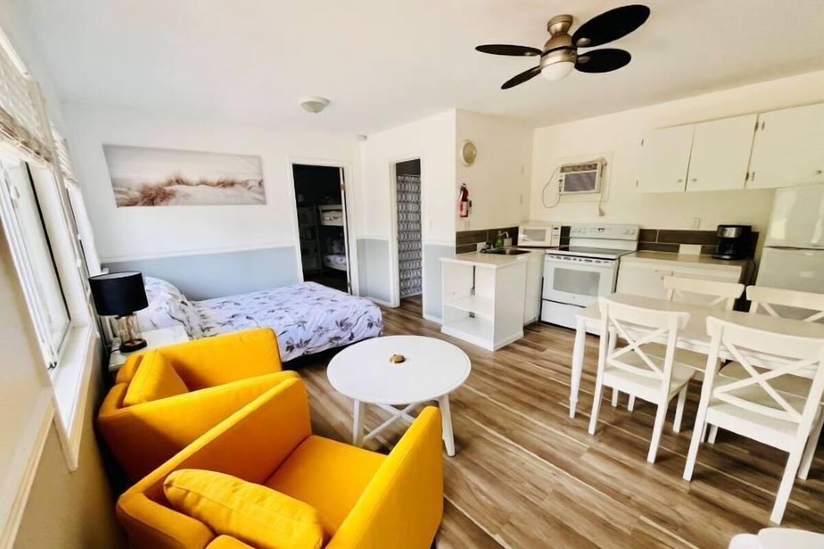 Un espacio habitable compacto con una cama, un sofá amarillo, una mesa de comedor con sillas, una kitchenette y un ventilador de techo.
