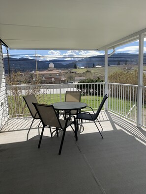 A covered patio with a table and chairs, offering a view of a rural landscape with fields and a church. - The Orchard at Oliver (Oliver)