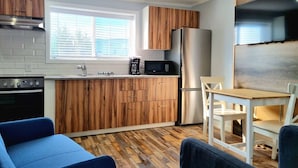 A compact kitchen with wooden cabinets, a stainless steel refrigerator, a microwave, and a coffee maker. There is a dining area with a table and chairs. - The Orchard at Oliver (Oliver)