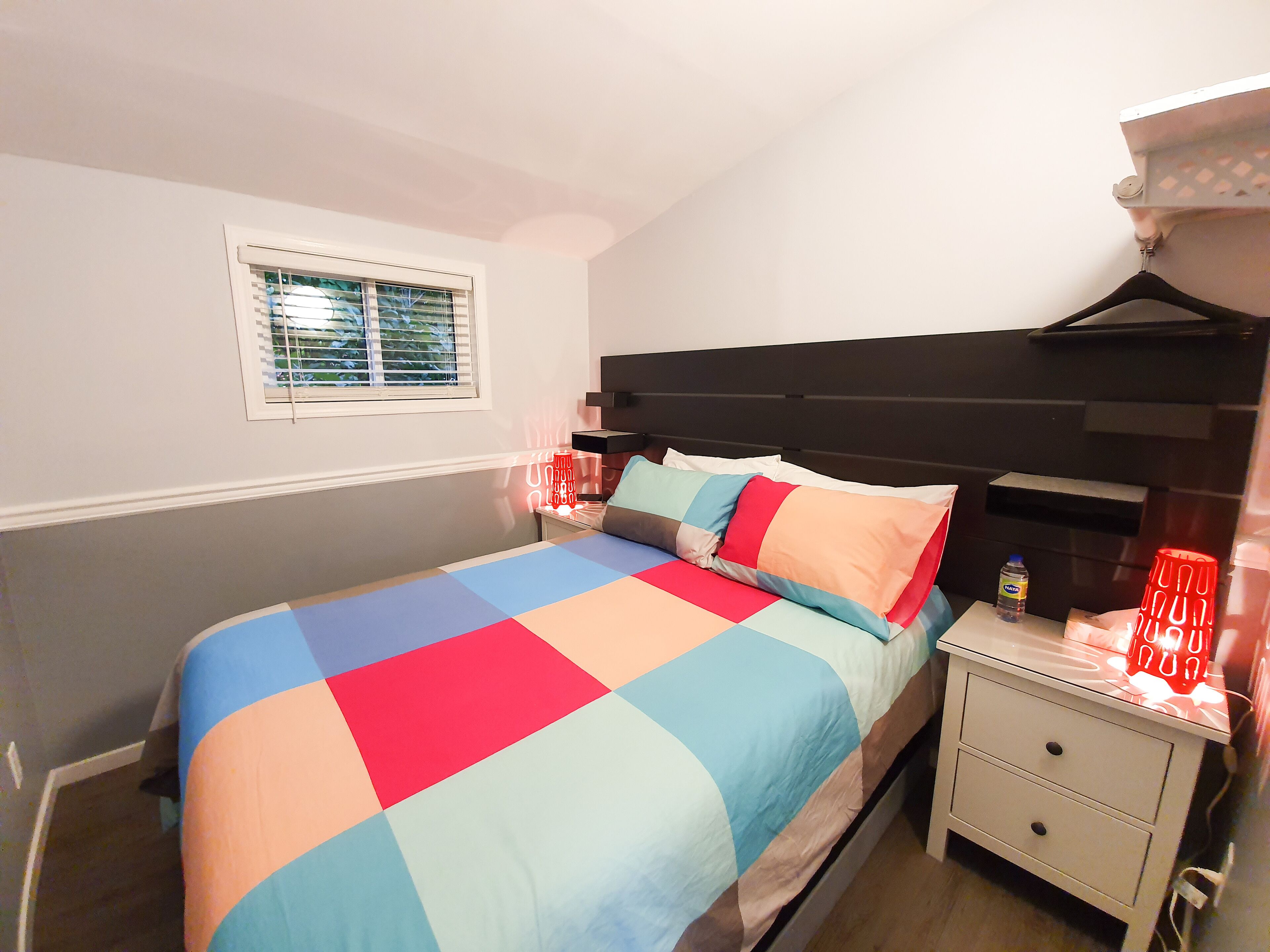 A bedroom with a bed, a nightstand, a window with blinds, and a wall-mounted shelf.