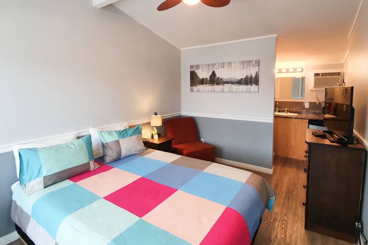 Un dormitorio con cama, mesita de noche, una silla roja, ventilador de techo, un cuadro, espejo y televisor.
