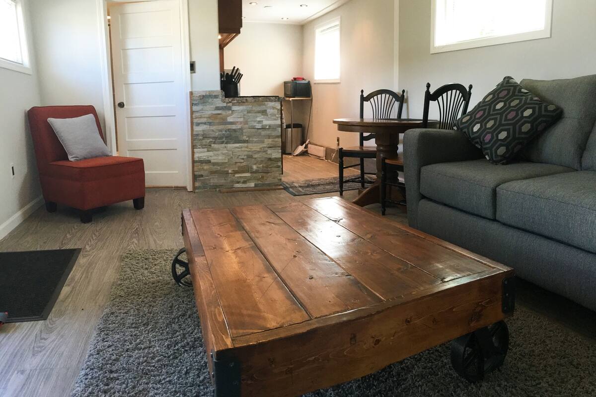 Una sala de estar con una silla roja, un sofá gris, una mesa de centro de madera, un muro de acento de piedra y una zona de comedor con una mesa y sillas.