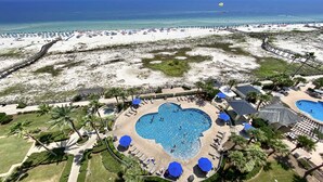 Vista aérea de un complejo turístico en la playa con una piscina grande con forma de corazón, rodeado de palmeras y tumbonas.