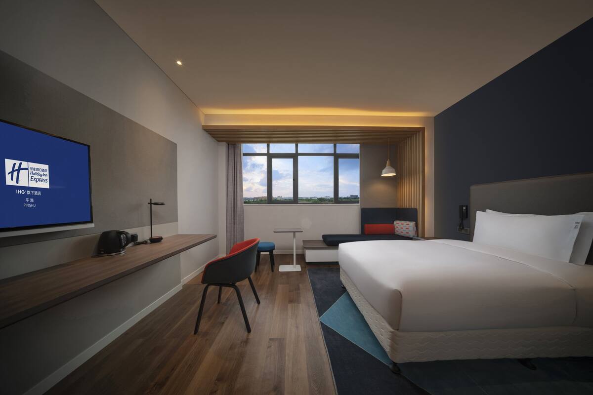 A modern hotel room with a large bed, a desk, a chair, a TV, and a window with a city view.