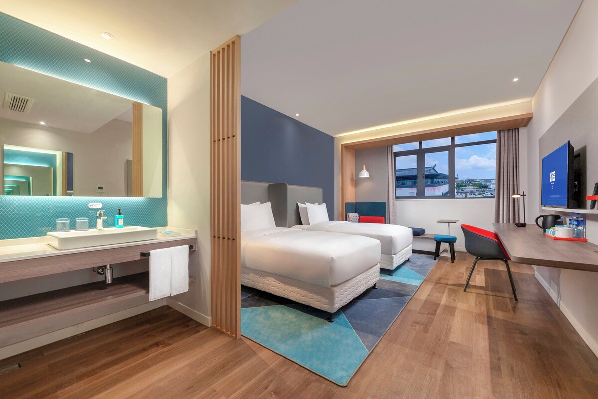 Ein modernes Hotelzimmer mit einem großen Bett, einem Schreibtisch, einem Stuhl, einem Fernseher und einem Badezimmer mit Spiegel und Waschbecken.