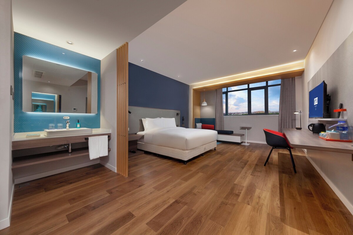 Ein modernes Hotelzimmer mit Bett, Schreibtisch, Stuhl, einem großen Fenster und einem Badezimmer mit Spiegel und Waschbecken.