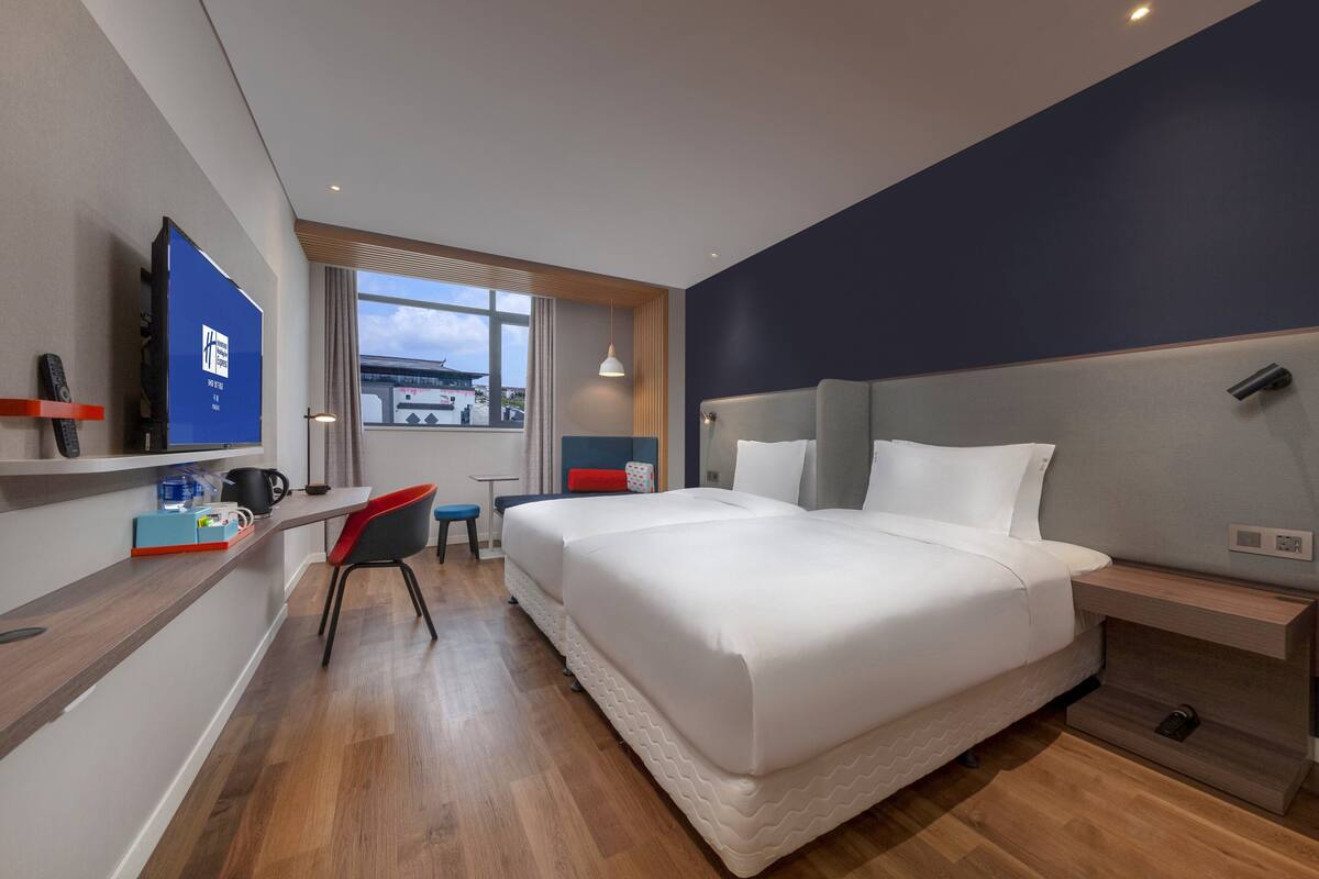 A modern hotel room with a large bed, a desk with a chair, a TV, and a window with a view of the outside.