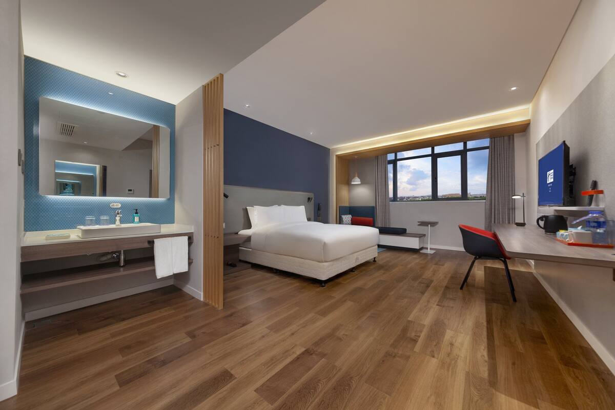 A modern hotel room with a wooden floor, a large bed, a desk with a chair, a TV, and a bathroom with a mirror and sink.