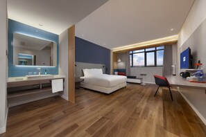 Ein modernes Hotelzimmer mit Holzboden, einem großen Bett, einem Schreibtisch mit Stuhl, einem Fernseher und einem Badezimmer mit Spiegel und Waschbecken.