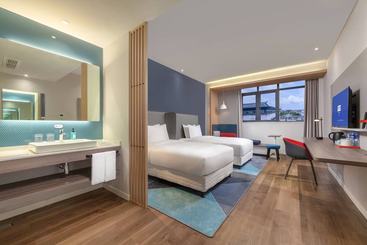 A modern hotel room with a large bed, a desk, a chair, a TV, and a bathroom with a sink and mirror.