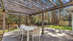 Una terraza de madera con un pérgola, una mesa blanca con sillas, y un patio cercado con árboles.