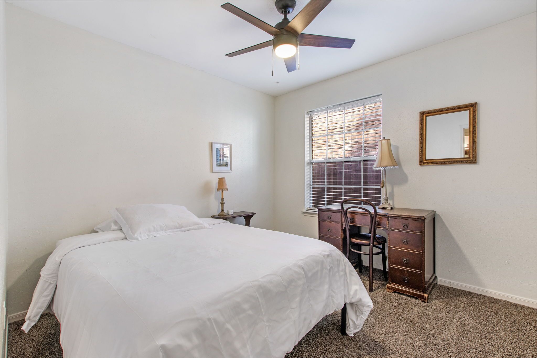 A bedroom with a bed, a dresser, a chair, a ceiling fan, a window with blinds, and a mirror on the wall.