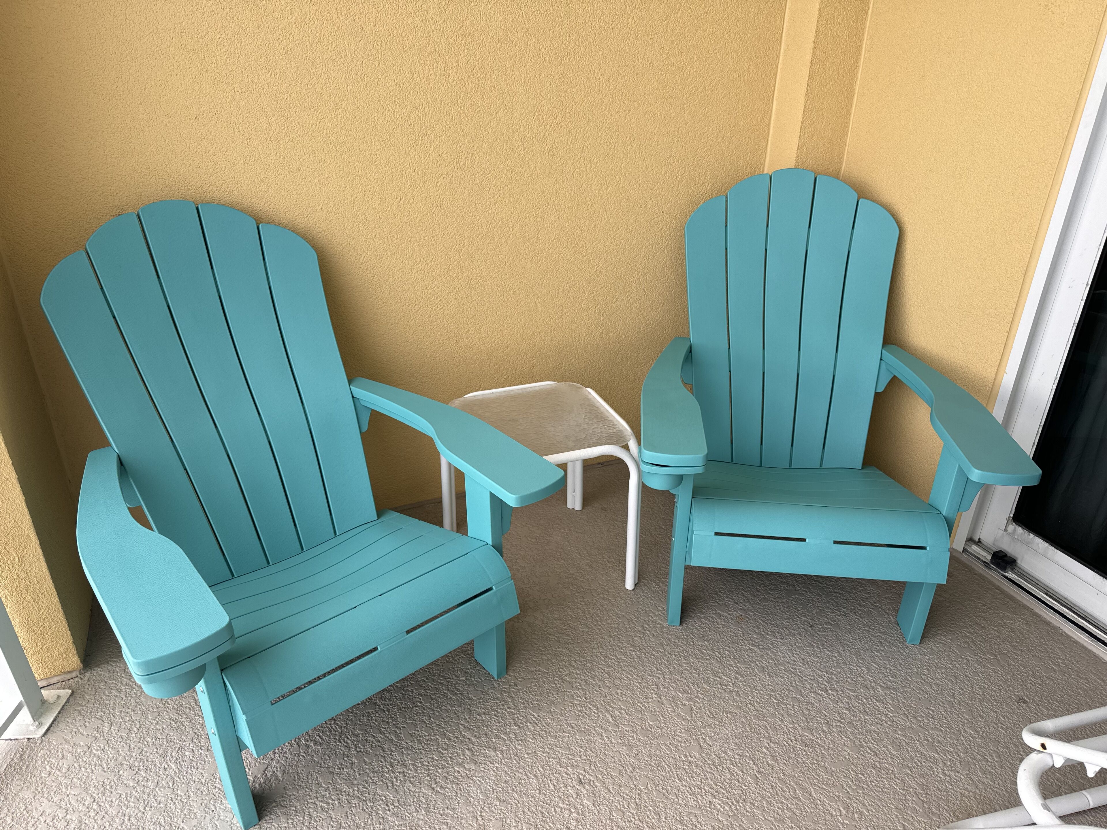 Two turquoise Adirondack chairs with white metal legs and a small white table between them, positioned on a textured beige carpet against a yellow wall.