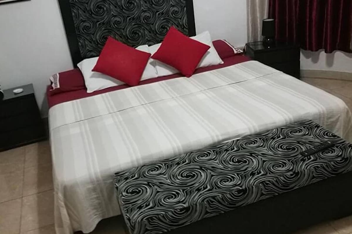 A bed with a black headboard, white and red bedding, and a black and white patterned bench.