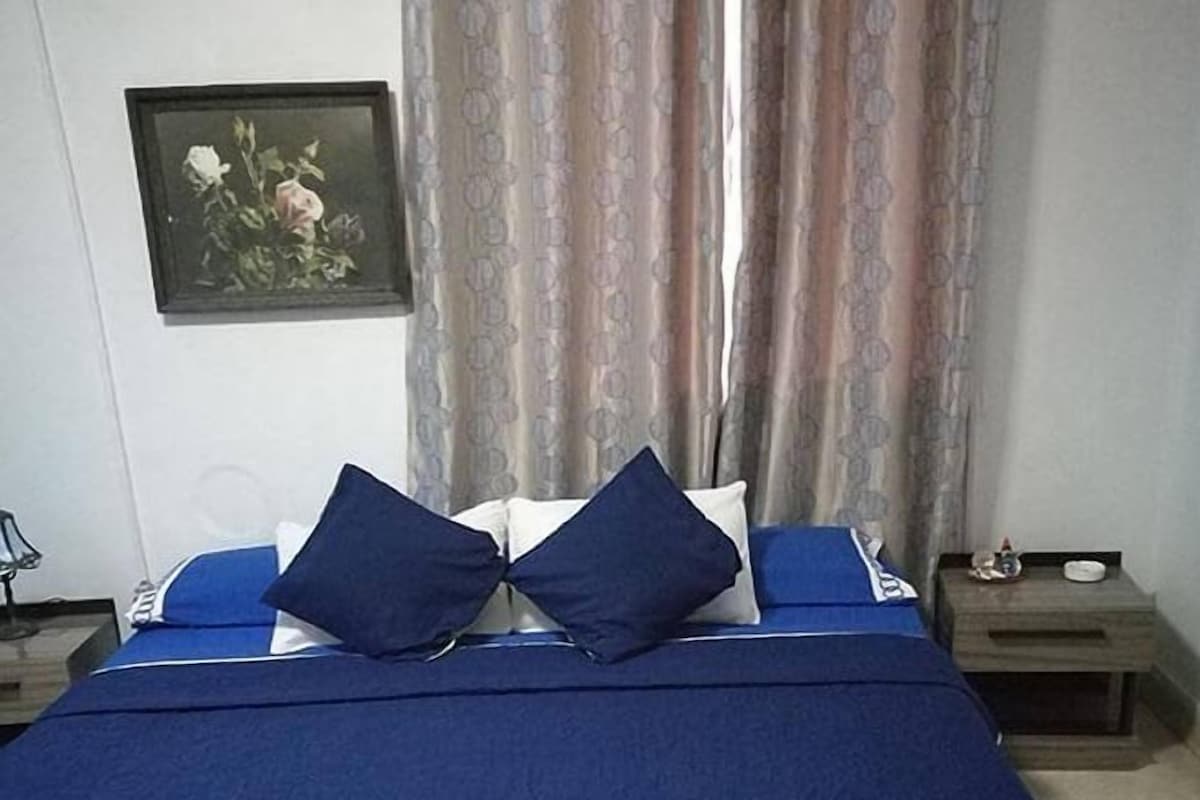 A bed with a blue bedspread and two pillows, a nightstand with a lamp, and a framed picture on the wall.