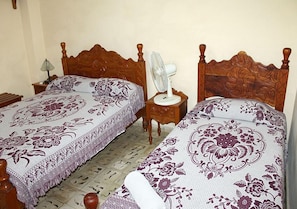 Ein Zimmer mit zwei hölzernen Betten, floraler Bettwäsche, einem Nachttisch mit Lampe und einem kleinen weißen Ventilator.