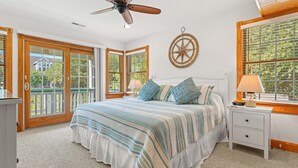 Une chambre à coucher comprenant un lit, une table de chevet, un ventilateur de plafond et une grande fenêtre avec des stores.