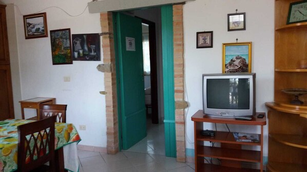 A living room with a television on a wooden stand, a dining area with a table and chairs, and a green door leading to another room.