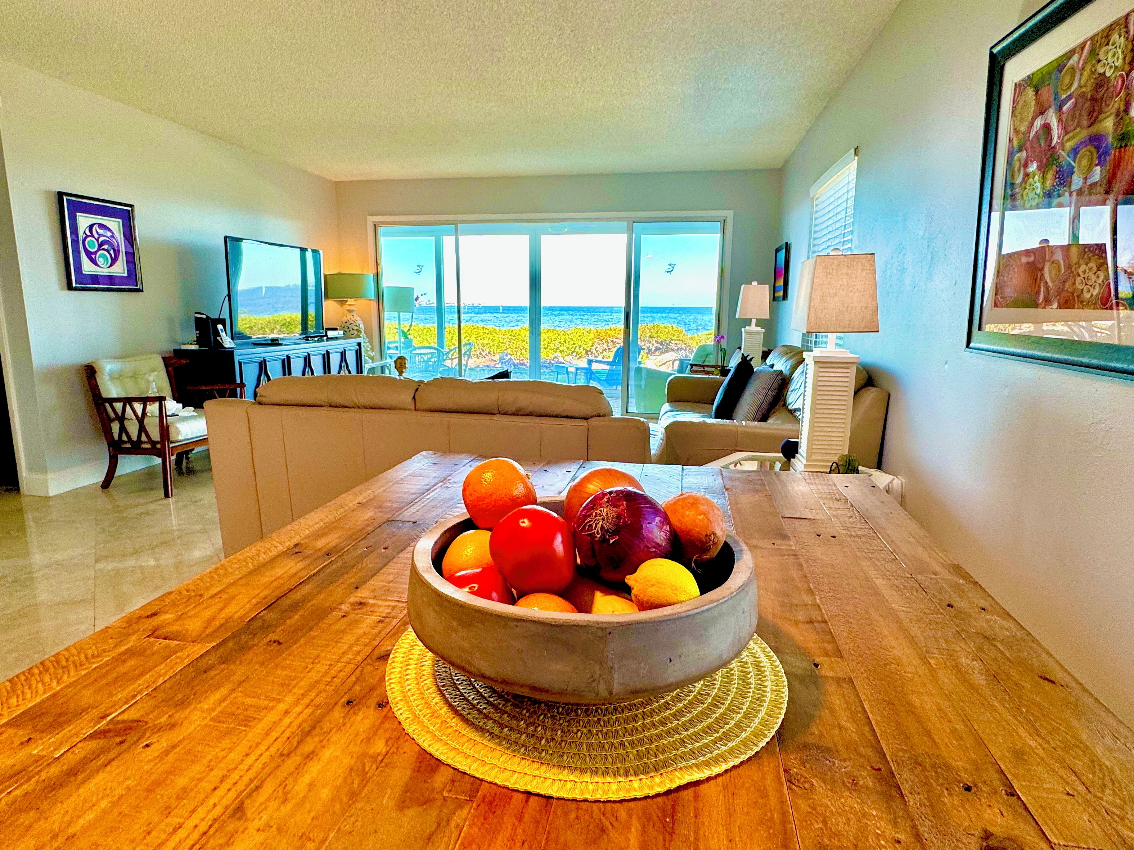 A living room with a wooden dining table, a bowl of fruit, a sofa, and a view of the ocean through large windows.