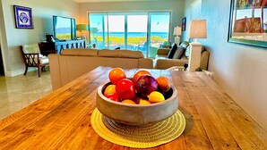 A living room with a wooden dining table, a bowl of fruit, a sofa, and a view of the ocean through large windows. - West Bay Cove 131 · Skyway Views · 30-Night Min (Holmes Beach)
