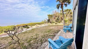 A patio with blue chairs, a view of the ocean, and a grassy area with a palm tree. - West Bay Cove 131 · Skyway Views · 30-Night Min (Holmes Beach)