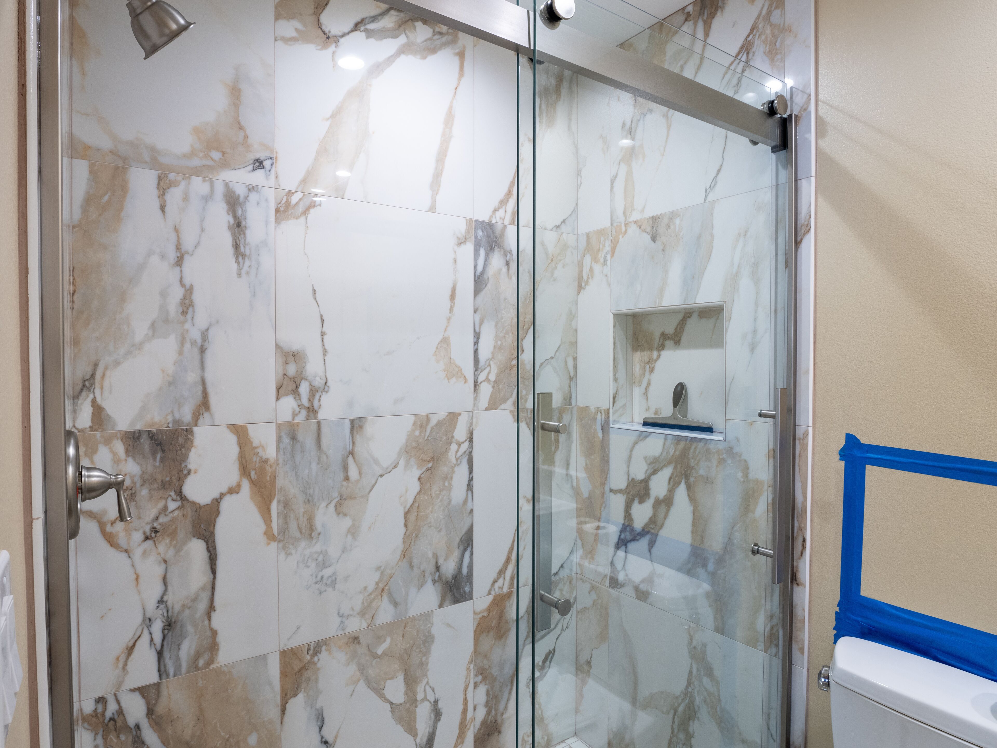 Un baño con baldosas de mármol, cabina de ducha de vidrio y un inodoro marcado con cinta de pintor azul.
