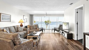 A modern living room with a large window offering a view of the sea, a sofa set, a coffee table, and a TV on a stand.