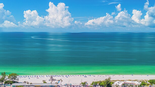 A beach with clear turquoise waters, a sandy shore, and a few buildings in the foreground.