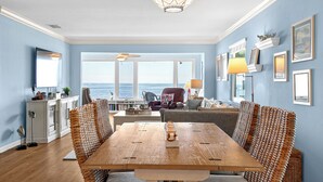 A dining area with a wooden table, wicker chairs, a ceiling fan, and a view of the ocean through large windows. - West Bay Cove 231 · Skyway Views · Sun Room (Holmes Beach)