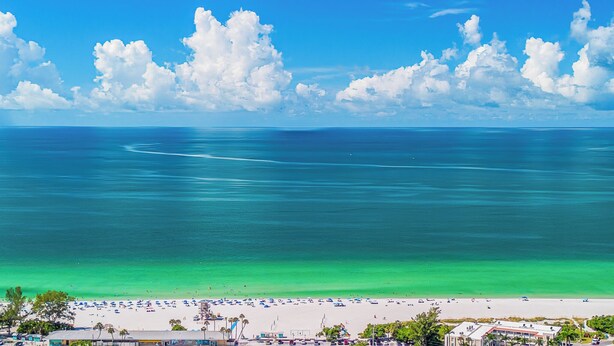 A beach with clear turquoise waters, a sandy shore, and a few buildings in the foreground. - West Bay Cove 231 · Skyway Views · Sun Room (Holmes Beach)