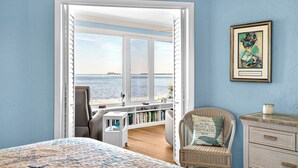 A room with a large window overlooking the sea, a wicker chair, a bookshelf, and a framed picture on the wall. - West Bay Cove 231 · Skyway Views · Sun Room (Holmes Beach)