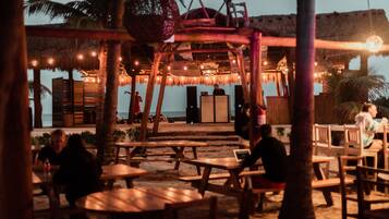 An outdoor seating area with wooden tables and chairs, a thatched-roof structure, and string lights.